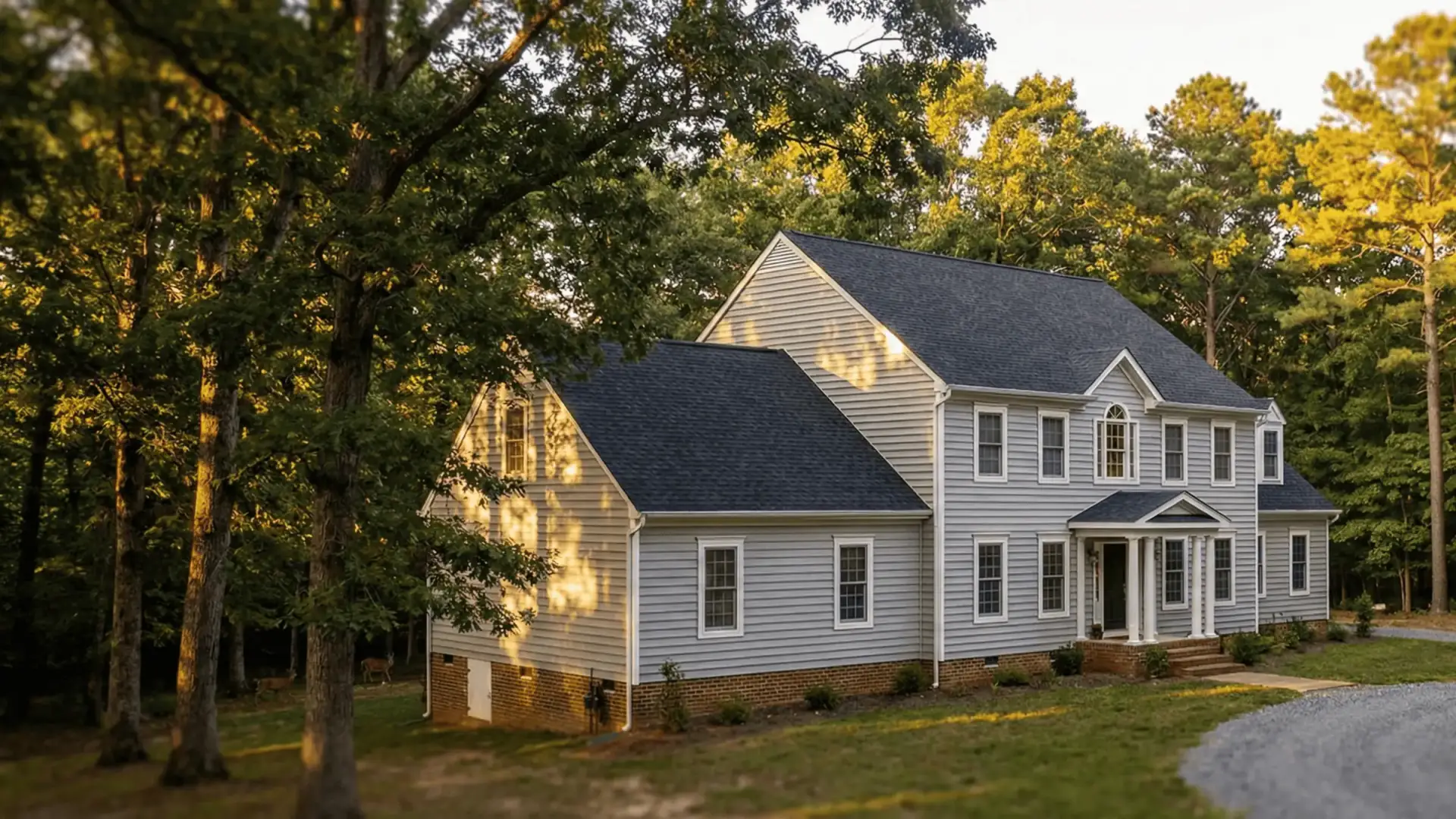 JDH Remodeling newly completed two-story colonial Huntingtown Maryland home with white vinyl siding, dark asphalt shingle roof, front portico, and replacement windows in a wooded setting